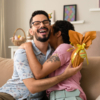 A couple sits on a couch, hugging each other joyfully, with an assortment of Easter chocolate gifts nearby