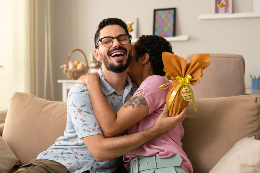 A couple sits on a couch, hugging each other joyfully, with an assortment of Easter chocolate gifts nearby