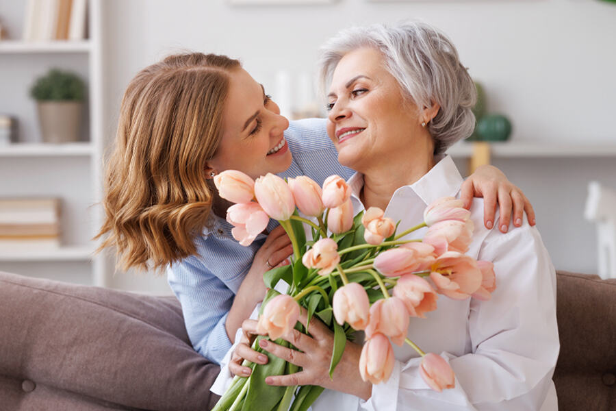 A woman embraces an older woman, presenting her with a bouquet of flowers in celebration of Mother's Day