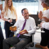 A group of colleagues gathers around a man presenting a gift box, celebrating Boss’s Day with thoughtful ideas