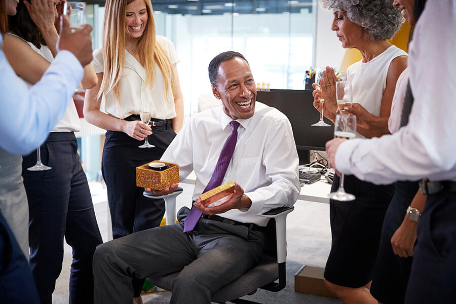 A group of colleagues gathers around a man presenting a gift box, celebrating Boss’s Day with thoughtful ideas